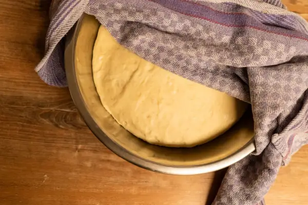 Ein goldgelber Pizzateig ruht in einer Schüssel, bedeckt mit einem karierten Tuch. Seine weiche, elastische pizzateig konsistenz verspricht eine köstliche Pizza.