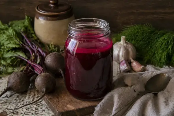 Słoik z kiszonym barszczem czerwonym, obok buraki, czosnek i koperek. Kiszony barszcz czerwony na co pomaga? Na odporność i trawienie!