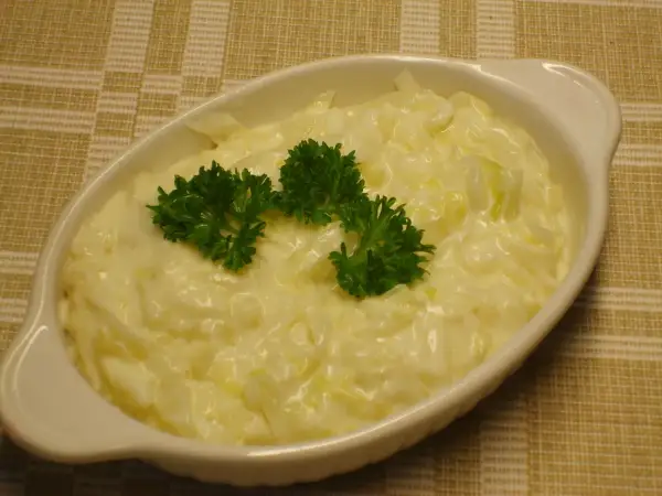 En vit oval skål med krämig stuvad vitkål, toppad med färsk persilja. Serveras på en rutig duk.