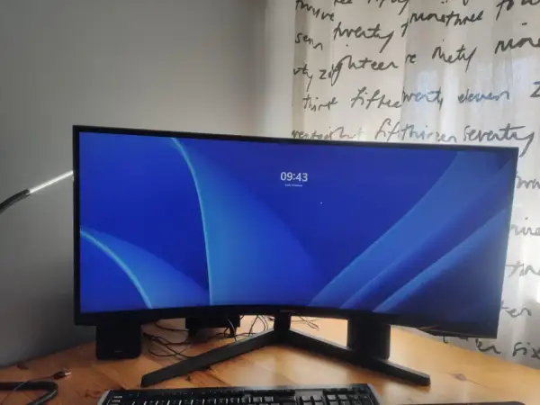 Zakrzywiony monitor 16:9 z niebieską tapetą i zegarem 09:43. Obok klawiatura i lampka.