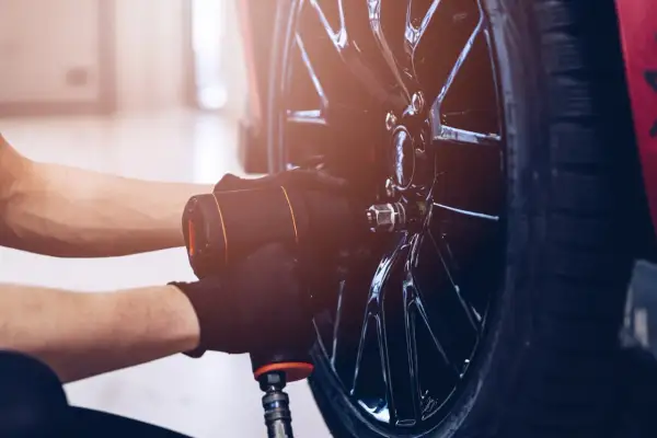 Mechanik w czarnych rękawiczkach używa klucza pneumatycznego do odkręcania śrub w feldze, przygotowując się do wymiany jednej opony.