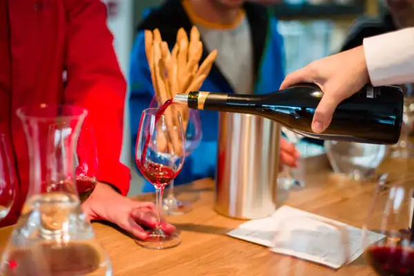 Ręka nalewa czerwone wino do kieliszka. W tle widać ludzi i przekąski.