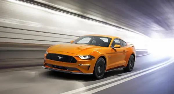 Un Mustang naranja acelera en un túnel, sus potentes sonidos de coches deportivos resonando.