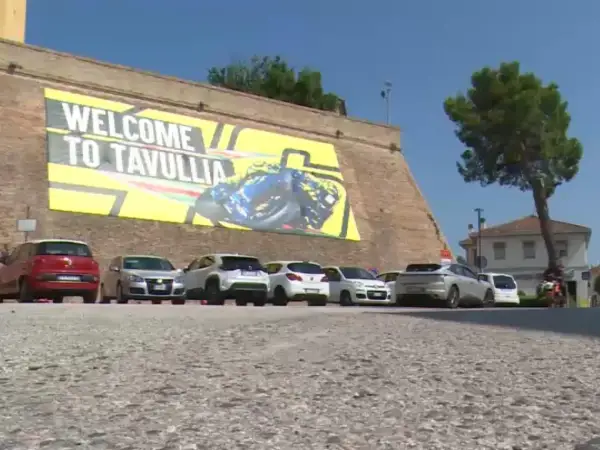 Benvenuti a Tavullia, la terra di Valentino Rossi! Un grande striscione giallo celebra il campione, con auto parcheggiate sotto.