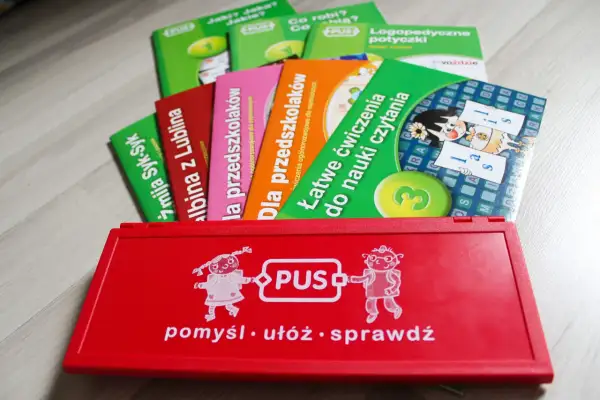 Czerwone pudełko z napisem "PUS" i rysunkami dzieci, obok kolorowe książeczki edukacyjne. Idealne do nauki i zabawy.