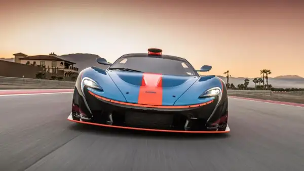 El McLaren P1, azul con franjas naranjas, acelera en una pista al atardecer, con palmeras y montañas de fondo.