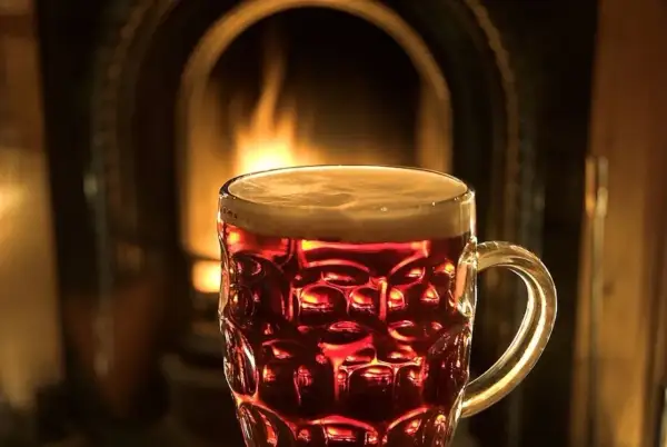 Bicchiere di birra ambrata con schiuma, davanti a un camino acceso. Un'atmosfera accogliente per gustare una birra calda.