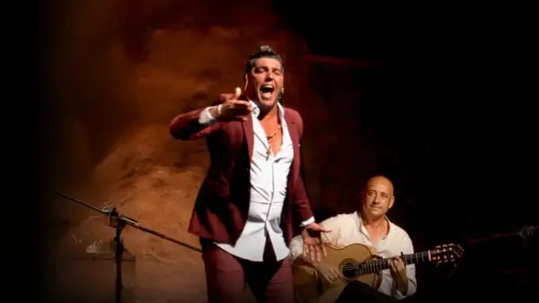 Un hombre con traje granate canta con pasión, su voz flamenco resuena mientras un guitarrista lo acompaña.