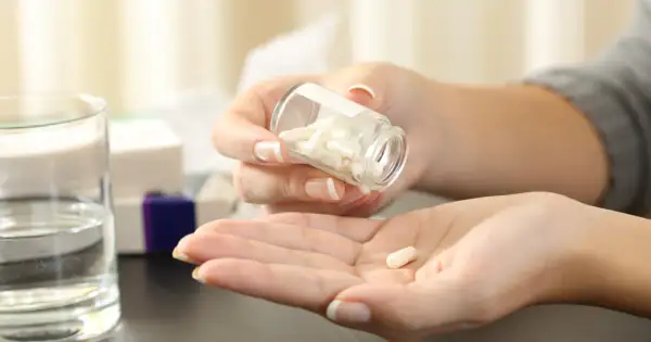 Kobieta wysypuje kapsułki z buteleczki na dłoń. Czy paracetamol jest przeciwzapalny? Obok stoi szklanka wody i pudełka leków.