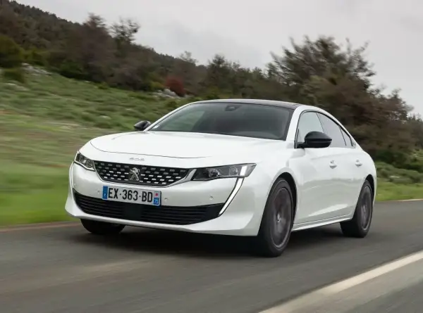 Biały Peugeot 508 jedzie po krętej drodze wśród zielonych wzgórz.
