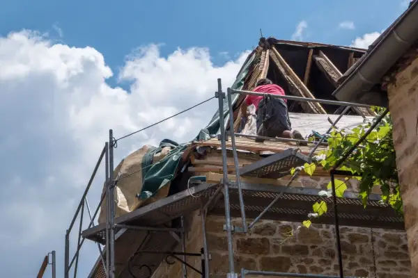 Un ouvrier travaille sur un toit en rénovation, une étape essentielle pour garantir ma prime logement décent. Le ciel bleu et les nuages blancs surplombent le chantier.