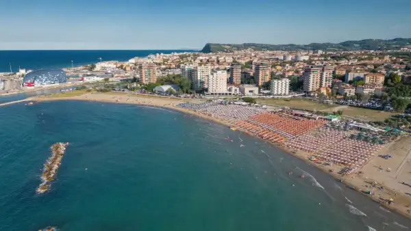 Vista aerea della Pesaro strand, con ombrelloni colorati, mare azzurro e la città sullo sfondo.