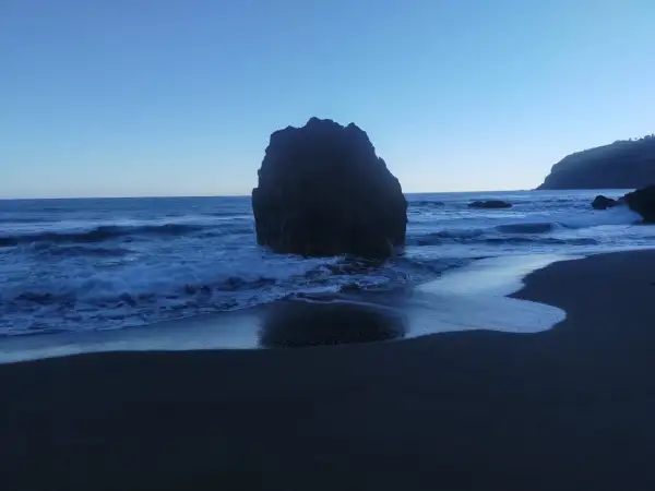 Olas rompen contra una gran roca en la playa los patos. El cielo azul claro se funde con el mar.