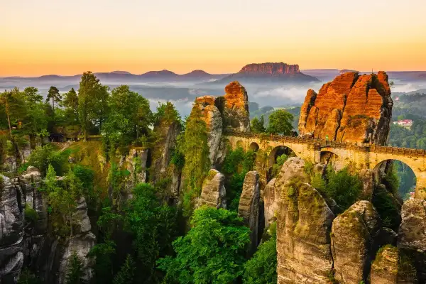 Un pont de pierre traverse des formations rocheuses spectaculaires, rappelant la Suisse bohémienne. La nature y est luxuriante, baignée par la lumière dorée du crépuscule.