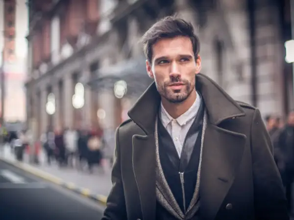 Ein Mann mit Bart trägt eine elegante, olivgrüne Jacke zum Anzug. Er blickt nach rechts, während er durch eine belebte Stadt geht.
