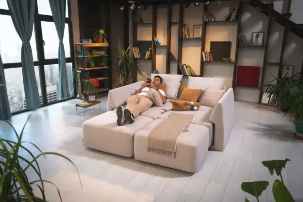Mann entspannt auf einem modularen Sofa, das wie ein **Bett als Sofa gestalten** wirkt. Gemütliche Kissen und eine Decke laden zum Verweilen ein.