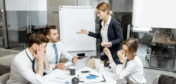 En kvinna gestikulerar mot en whiteboard medan kollegor ser utmattade ut. En känsla av passiv aggressiv kommunikation genomsyrar mötet.
