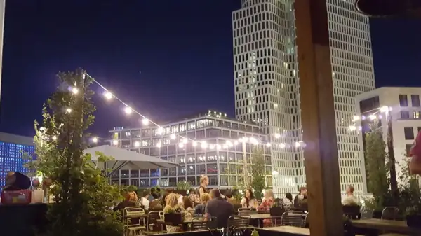 Abendstimmung auf einer Dachterrasse in Berlin. Lichterketten beleuchten Tische, an denen Gäste sitzen. Im Hintergrund ragen moderne Gebäude auf, die an den Spreegold Bikini erinnern.