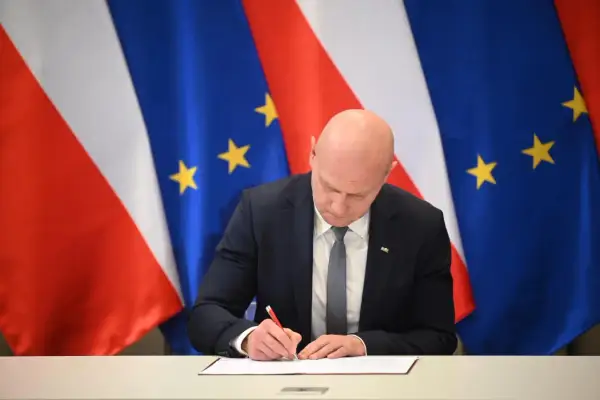 Łysy mężczyzna w garniturze podpisuje dokument na tle flag Polski i UE. Spodziewaj się niespodziewanego, bo nigdy nie wiadomo, co przyniesie przyszłość.