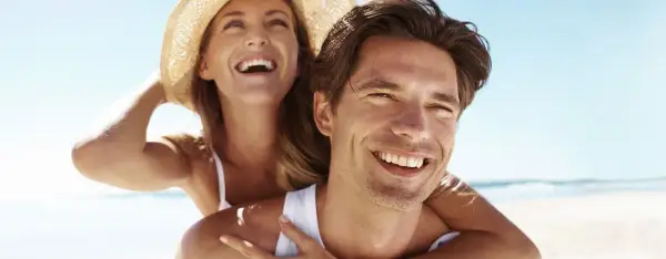 Glückliches Paar am Strand, die Frau lacht und trägt einen Hut. Selbst im Schatten wird man braun, wenn man die Sonne genießt.