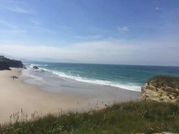 Un paseo solitario por las **playas de Lugo**, con el mar turquesa rompiendo suavemente en la arena dorada y acantilados rocosos.