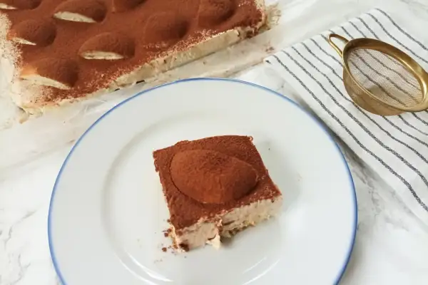 Pyszne ciasto cappuccino z mascarpone, posypane kakao, czeka na talerzu. Obok całe ciasto i sitko do posypywania.