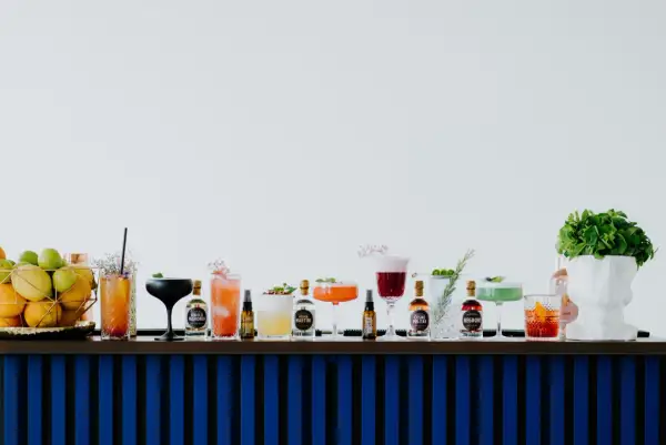 Rząd kolorowych drinków i butelek z alkoholami na barze, obok koszyka z owocami i białej rzeźby głowy.