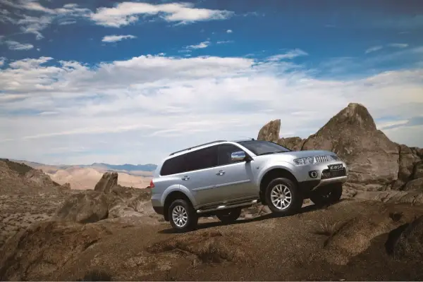 Srebrny Mitsubishi Pajero Sport pokonuje skalisty teren na tle pustynnego krajobrazu i błękitnego nieba z chmurami.