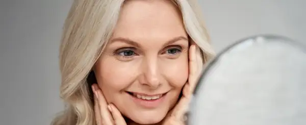 Uśmiechnięta 40-latka z blond włosami, patrząca w lustro. Delikatny makijaż na wesele podkreśla jej urodę.