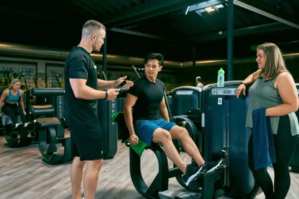 Trainer erklärt Mann die richtige Ausführung am Gerät für den erector spinae. Frau schaut zu.