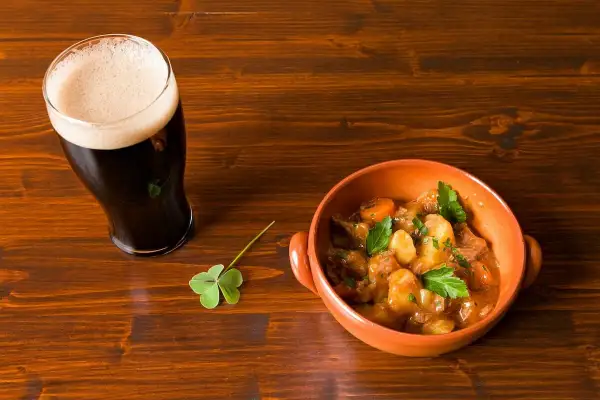 Un bicchiere di birra stout scura con schiuma cremosa accanto a una ciotola di stufato irlandese e un quadrifoglio su un tavolo di legno.