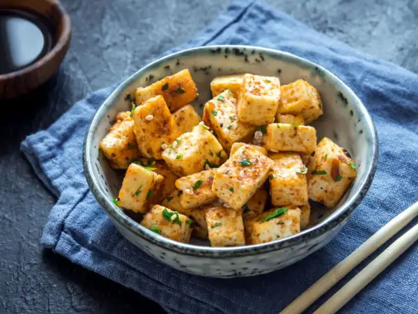 Usmażone kostki tofu, które jest zrobione z ziaren soi, w misce z sezamem i ziołami. Obok pałeczki i sos sojowy.