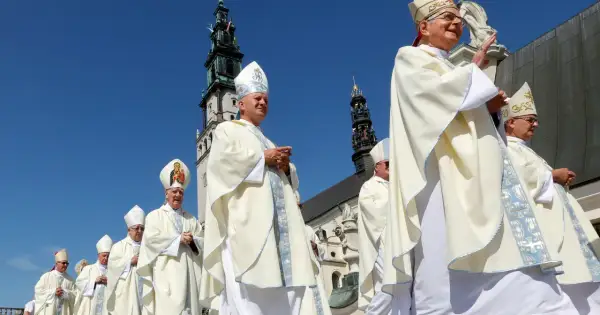 Biskup w białej szacie i mitrze z Matką Boską, na tle wieży kościelnej.