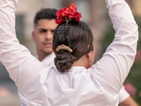 Peinados para bailar flamenco: moño bajo adornado con flores rojas y peineta dorada. Un hombre con camisa blanca al fondo.
