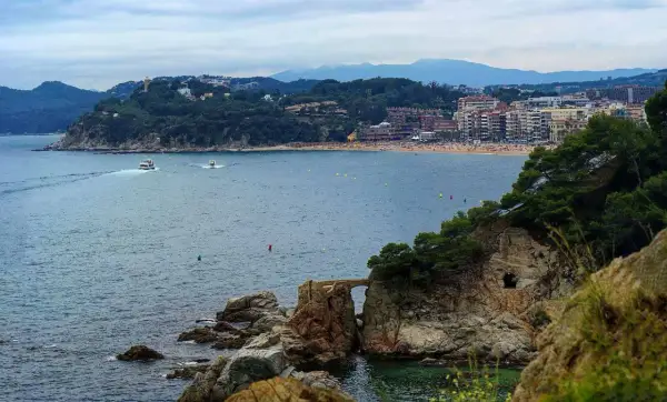 Widok na zatokę z łodziami i plażą. W oddali widać miasto i góry. Pogoda na Costa Brava zapowiada się na spokojną.