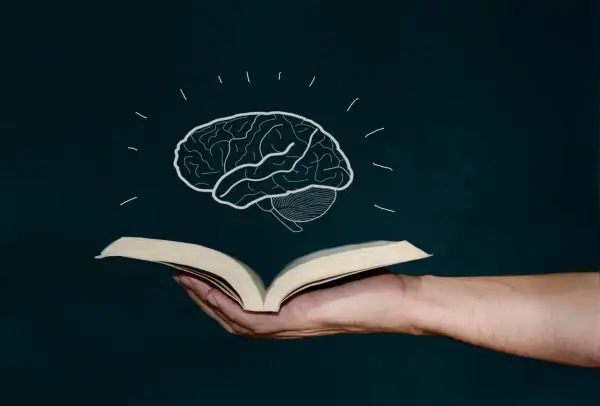 Mano sostiene libro abierto, de él surge un cerebro dibujado, símbolo de la lectura global y el conocimiento.