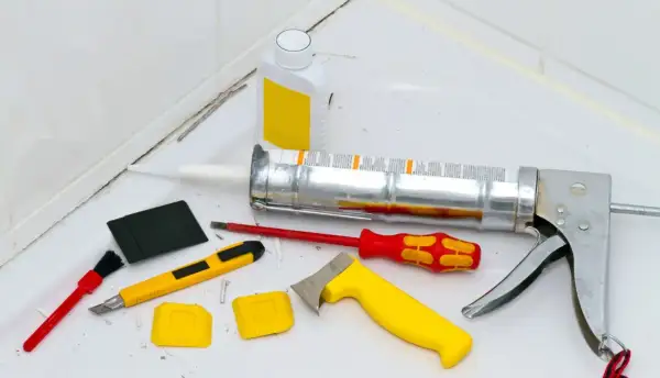 Werkzeuge zum Silikon von Fliesen entfernen: Kartuschenpresse, Cutter, Schaber, Bürste und eine Flasche mit Reiniger.
