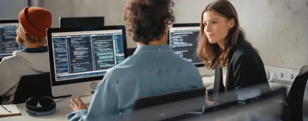 Dwóch programistów przy komputerach, jeden w pomarańczowej czapce, drugi w dżinsowej koszuli. Rozmawiają o tym, jak się przebranżowić na IT.