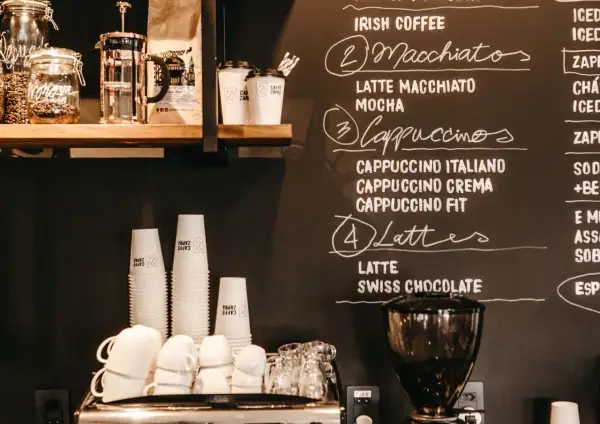 Tafel mit Kaffeebar Ideen: Macchiatos, Cappuccinos, Lattes und mehr. Stapel von Pappbechern und Espressomaschine im Vordergrund.