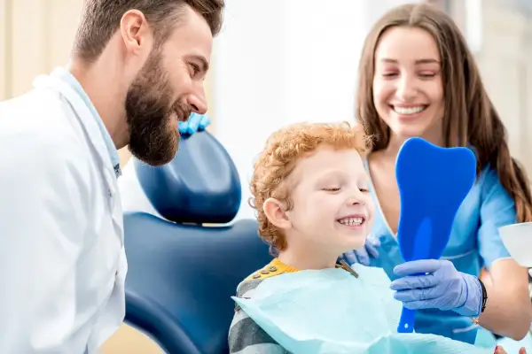 Uśmiechnięty chłopiec ogląda swoje zęby w lusterku w kształcie zęba, podczas gdy dentysta i asystentka patrzą z aprobatą. Czy fluoryzacja jest szkodliwa? Nie, to zabieg chroniący zęby.