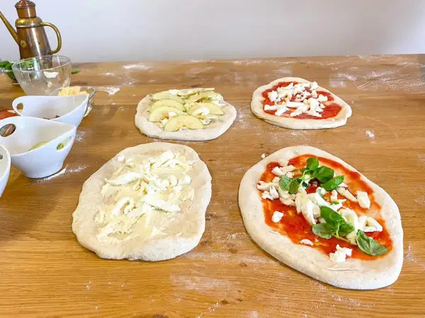 Vier rohe Pinsa-Teige auf einem Holztisch, teils belegt mit Tomatensoße, Mozzarella, Äpfeln und Basilikum.