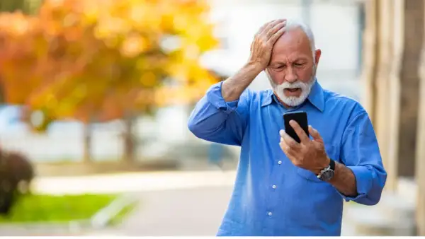 Anziano con mano sulla testa, guarda il telefono con espressione preoccupata, forse per la paura di parlare al telefono.