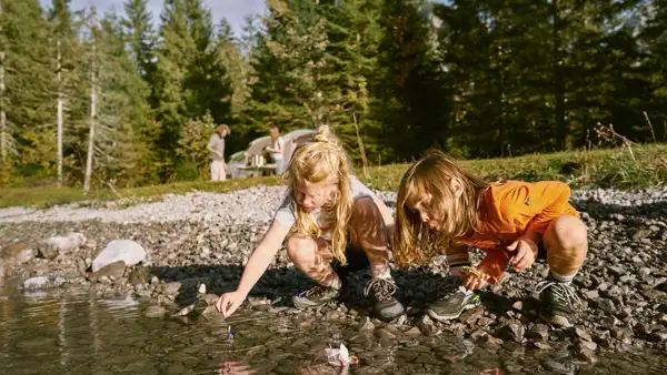 Två barn leker med pappersbåtar vid en sjö. I bakgrunden syns ett tält och träd. En härlig 5 kamp fest i naturen.