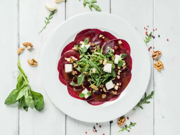 Rote Bete Carpaccio mit Rucola, Walnüssen und Feta auf einem weißen Teller, garniert mit frischem Basilikum.