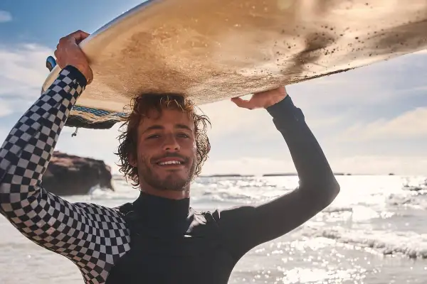 Lächelnder Surfer mit lockiger Surfer-Frisur trägt Neoprenanzug und hält Surfbrett über dem Kopf.