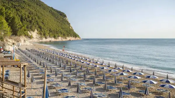 Filettata di ombrelloni e lettini sulla spiaggia di San Michele, con il mare azzurro e la collina verde sullo sfondo.