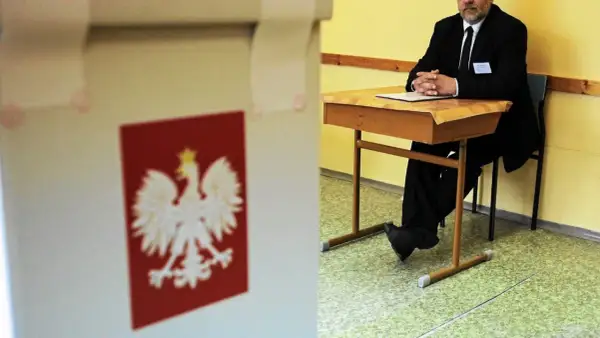 Mąż zaufania w garniturze siedzi przy biurku w lokalu wyborczym, obok urny z godłem Polski.