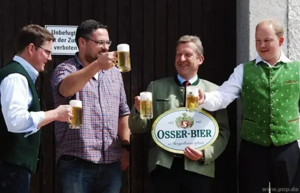 Vier Männer stoßen mit Biergläsern an. Einer hält ein Schild der Osser Brauerei.