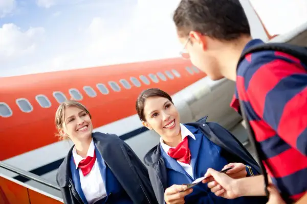 Zwei lächelnde Flugbegleiterinnen im blauen Anzug mit roten Halstüchern prüfen eine Bordkarte vor einem Flugzeug. Die Ausbildung zur Flugbegleiterin und das Gehalt sind attraktiv.