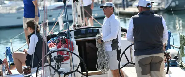 Equipaggio barca a vela pronto per la regata. Un uomo tiene una cima, una donna è seduta, mentre un altro uomo è al timone.